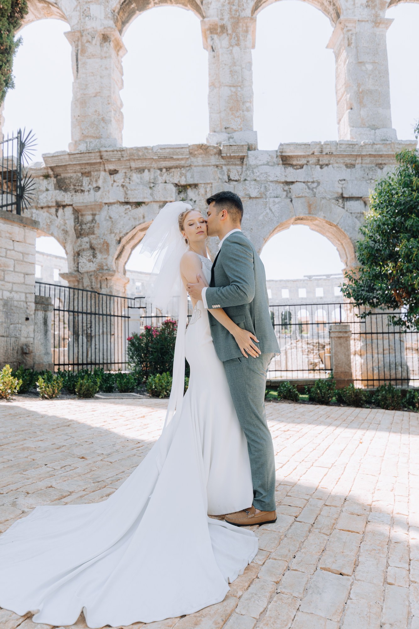 Elena und Tyson in Hochzeitskleidung vor den hellen Bögen des Amphitheaters in Pula